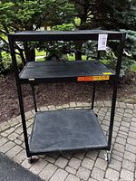 Front angled view showing three shelves and open frame of black metal AV cart with wheels on paved surface outdoors.