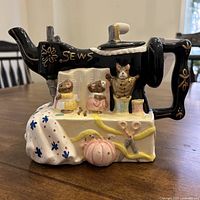 Photo showing ceramic teapot shaped like a vintage sewing machine with a design of three mice and sewing accessories on the front