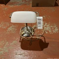 Front angled view of the hardwired brass-colored metal and glass bankers lamp showing white glass shade, circular metal base with decorative perforations, pull chain, and attached cord on concrete floor.