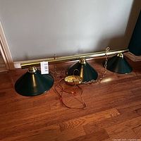 Full view of the green and brass billiard light fixture showing three conical green metal shades attached to a horizontal brass-colored bar, with hanging chain and wiring on wooden floor.