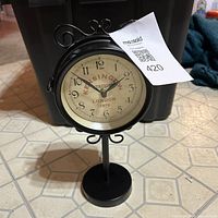 Front view of black metal double-sided desk clock, showing cream face with 'Kensington Station London 1879' logo and black numerals.