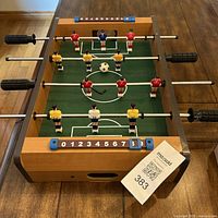 Top front view of mini foosball table showing wooden frame, six players per team, manual slider scoring, and included foosball ball.
