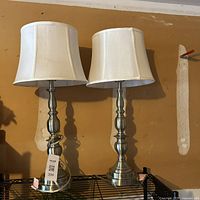 Front view of two modern table lamps with silver metal bases and white fabric shades, showing the overall design and size.