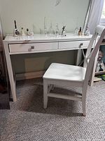 Front and side view of white wood desk and chair in carpeted room with decorative glass and ceramic items on desk.