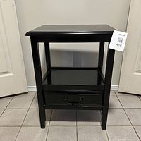 Front view of the black wood side table showing drawer with handle and open lower shelf, standing on tiled floor.