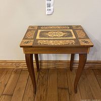 Front angled view of wooden table showing the inlaid floral and geometric pattern on the top surface and curved legs.