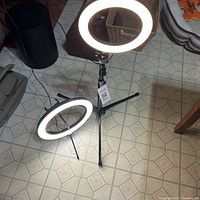 Photo showing two ring lights illuminated on tripod stands, one medium-sized and one smaller with phone holder visible, all on tiled floor.