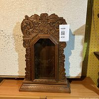 Front view of ornate carved wood clock case with glass door and decorative engraving, displaying slight damage on upper right corner.