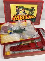 Open box of vintage Meccano set showing red and green metallic strips, parts, and wheels wrapped in bubble wrap inside original box with artwork depicting children building.