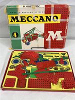 Open box showing yellow plastic insert tray filled with steel Meccano parts including red perforated plates, green perforated strips, wheels, pulleys, and brackets.