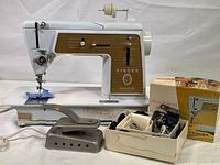 Full view of the Singer Model 603 sewing machine on the table with attached fabric and the box of attachments and the vintage iron beside it.