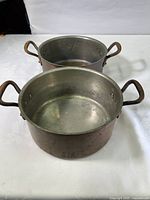 Two vintage copper and steel saucepans with metal side handles on white surface, showing round shape and patina on copper exteriors.
