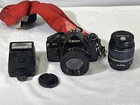 Photo showing Canon A-1 camera with FD 50mm lens attached, extra 18-55mm lens, Canon flash unit, lens cap and red camera strap laid out on white surface.