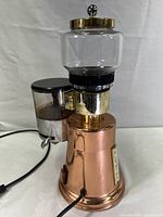 Full unit view showing copper base, glass bean hopper, stainless steel grinder mechanism, plastic lid, and power cord