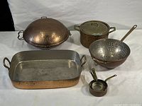Photo showing entire collection of vintage copper and brass cookware items on white background, including dome lid, rectangular baking dish, pot, colander, and nested measuring cups.