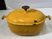 Top and side view of orange/yellow oval cast iron Dutch oven with lid on showing wear and chip on lid.