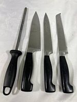 Four different cutting utensils including sharpening steel, chef's knife, smaller chef's knife, and serrated knife placed on cloth background.