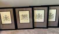 Four framed coat of arms art prints placed on floor, black frames with linen matting and visible reflections.