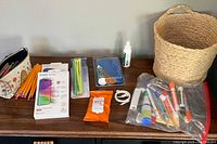 Photo showing pencils, colorful cord ties, screen protector packages, cleaning wipes, small spray bottle, zip pouch with pens, and dry erase markers in a clear plastic bag, placed next to a woven basket.