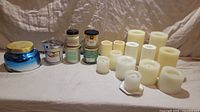 Overview of 13 battery operated candles in varying heights and 6 scented burnable candles in glass jars