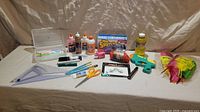 Shows a group of craft and art supplies including glues, markers, feathers, scissors, paint brushes, reusable bag, and a pink cow stress ball arranged on a white backcloth.