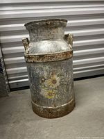Full view of the tall metal container showing aging, rust, and the floral design on its side, placed on a concrete floor against a metal backdrop.