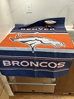 Photo showing the Denver Broncos yard flag laid out flat with the mini crock placed on top, highlighting the flag's colors and logo.