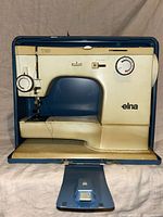 Elna sewing machine inside a blue protective case, showing the machine's overall front and side profile including the brand, stitching arm, and base.