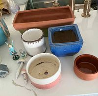 Photo of assorted flower pots including rectangular terracotta pot, white pot, blue square pot, reddish-brown round pot, and white pot with red checkered pattern alongside small figurines on white countertop.