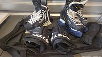 View of two pairs of boys' hockey skates, black Tracks size 8.5 and black Bauer size 10.5 with yellow and white accents, along with black Easton gloves, and a black duffle bag beneath