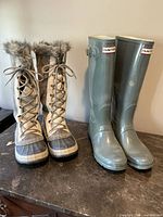 Two pairs of boots displayed side by side: Hunter dark green rubber boots with buckle and Sorel beige winter boots with faux fur lining and stitched suede upper.
