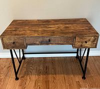 Front view of industrial wood and metal desk with three drawers, showing wood grain and metal legs.