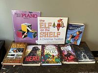 Photo showing the front covers of 8 kids books arranged side by side on the floor against a wall. Titles visible include Bastien Piano Basics Level 1, The Elf on the Shelf: A Christmas Tradition, The Littlest Ghost, The Story of Roy Rogers and Dale Evans: Happy Trails, Hero, Wonderful Wizard of Oz, Used of Magic, and Dark is Rising.