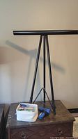 Large black aluminum flip top stand with tripod base and horizontal top bar placed on a table next to a container of craft supplies and blue hot glue gun.