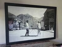 Black and white vintage style photo print of two men lifting a table with mountains in background, framed in black.