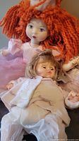 Photo showing two of the dolls, one with bright orange yarn hair and blue eyes in a pink dress, and one smaller doll with short brown hair and white outfit.