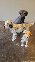 Three dog figurines arranged on a surface: one cast iron black dog, a white and black ceramic dog, and a smaller white and brown ceramic dog with a bone.