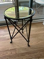 Side angle front view of the round dark bronze metal framed side table with glass top, showing the crossbar supports and legs.