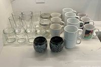 Overview photo showing all items: six hexagonal glass jars with lids, four round jars with lids, six tall clear drinking glasses, seven white ceramic mugs including two with floral designs, two black decorative candle holders.