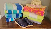 Photo of four folded beach towels with colorful stripes and floral patterns, a natural straw hat with a pink flower, and a pair of navy blue patent leather clogs for kids.