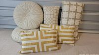 Six accent pillows displayed against a gray background; includes two round cream-colored pillows and four rectangular pillows with neutral geometric patterns in beige and mustard