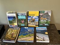 Eight outdoor and travel books arranged on table showing titles like Sri Lanka, Colorado hikes, bike maps, children activities, and Colorado 14ers.