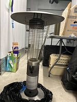 Full view of the outdoor heat lamp with metal top, protective cage around heating element, and base, next to cleaning supplies and storage boxes.