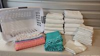Overall view of sheets, pillowcases, colorful throw, tablecloth, and laundry basket. Sheets and pillowcases are folded in stacks, mostly white. Laundry basket is white plastic missing one handle. Colorful throw patterned with fringe detail.