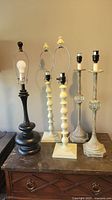 Six traditional style table lamps, including black and cream wooden bases and metal hardware without shades, shown on wooden surface
