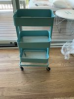 Full view of three-tier teal mesh cart on black casters, standing on wooden floor near glass door.