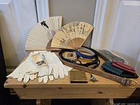 Photo of lady's accessories including two decorative fans, four belts, four gloves, three wallets/cardholders, a small gold-toned case, and assorted pins on a wooden surface.