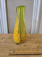 Front and side view of a signed Murano style glass vase with yellow, orange, and red patterns measuring about 7 inches tall, sitting on a wooden surface with ruler for scale.