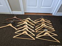 15 wooden hangers on carpet floor with door in background. Hangers in natural and dark wood finishes, one with clip attachment.