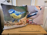 Two decorative throw pillows with bird images on a wooden surface against a white wall.